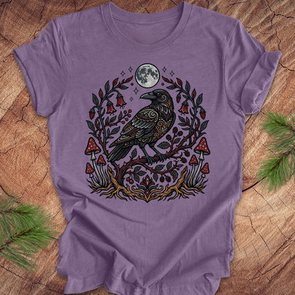 A lavender purple short-sleeve t-shirt with a detailed graphic of a black raven perched on a branch with red mushrooms, green leaves, and a full moon, laid flat on a rustic wood background with pine needles.