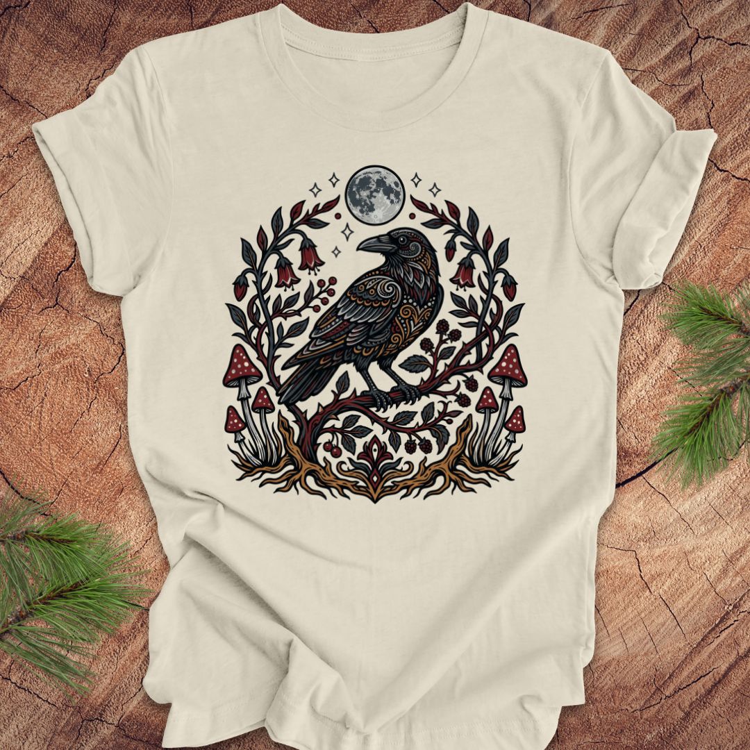 Beige t-shirt with a raven and nature-themed design on a wooden surface.