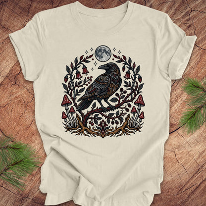 Beige t-shirt with a raven and nature-themed design on a wooden surface.