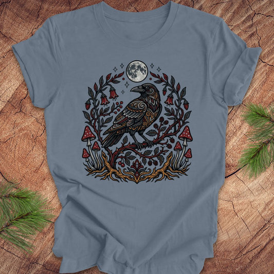 Steel blue t-shirt with a raven and nature-themed design on a wooden surface.