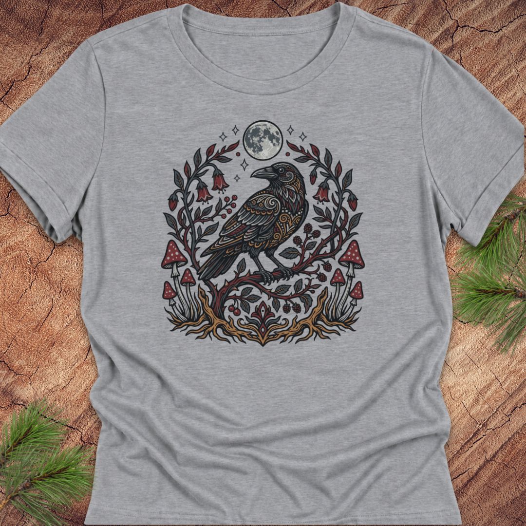 A heather gray short-sleeve t-shirt with a detailed graphic of a raven perched on a branch with red mushrooms, green leaves, and a full moon, laid flat on a rustic wood background with pine needles.