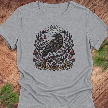 A heather gray short-sleeve t-shirt with a detailed graphic of a raven perched on a branch with red mushrooms, green leaves, and a full moon, laid flat on a rustic wood background with pine needles.