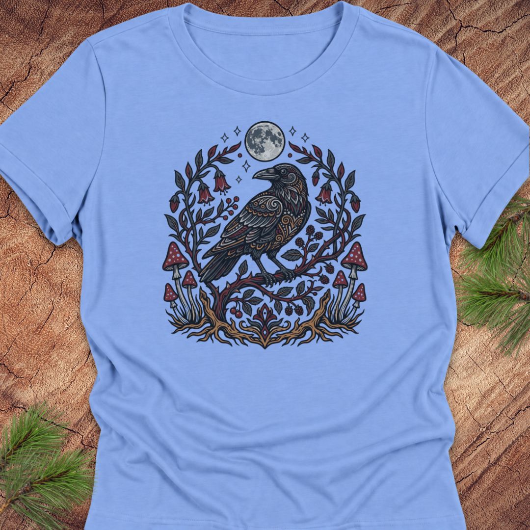A light blue short-sleeve crewneck t-shirt featuring a detailed graphic of a raven perched on a branch with red mushrooms, green leaves, and a full moon, laid flat on a rustic wood background with pine needles.