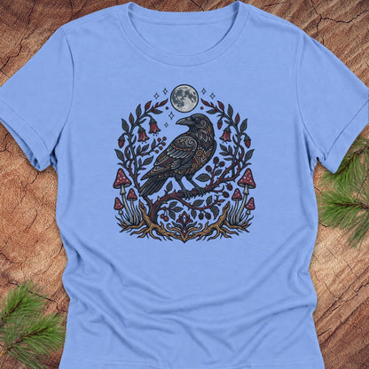A light blue short-sleeve crewneck t-shirt featuring a detailed graphic of a raven perched on a branch with red mushrooms, green leaves, and a full moon, laid flat on a rustic wood background with pine needles.