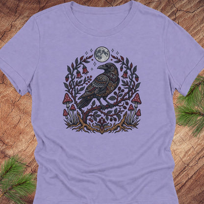 A lavender purple t-shirt featuring a detailed graphic of a raven perched on a branch with red mushrooms, green leaves, and a full moon, laid flat on a rustic wood background with pine needles.