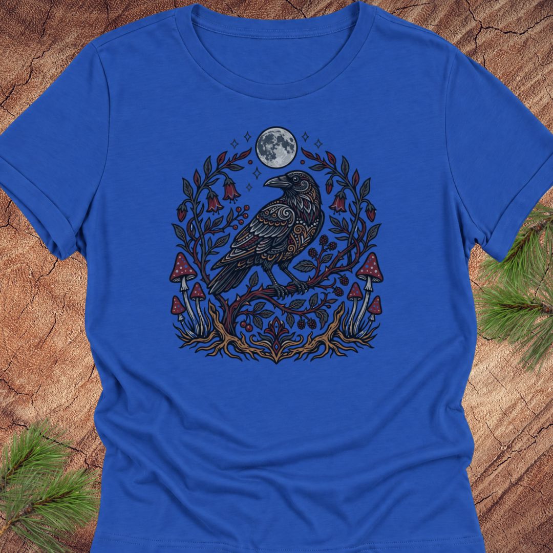 A royal blue short-sleeve crewneck t-shirt featuring a detailed graphic of a raven with intricate patterns, surrounded by a circle of mistletoe, bells, mushrooms, and a full moon, laid flat on a rustic wood background with pine branches.