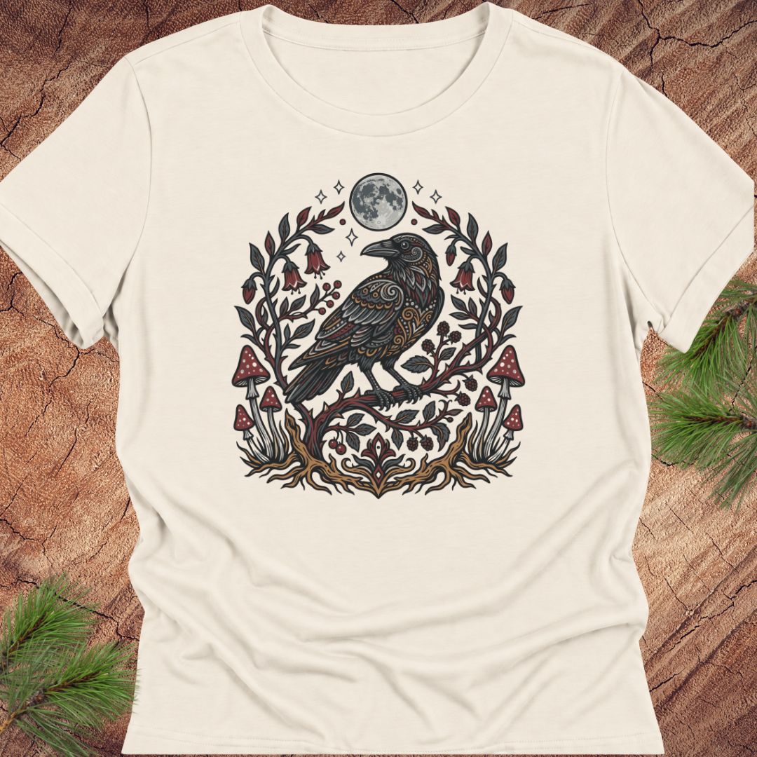 An off-white short-sleeve t-shirt with a detailed graphic of a black raven surrounded by a floral wreath, red mushrooms, and a full moon, displayed flat on a rustic wood background with pine needles.