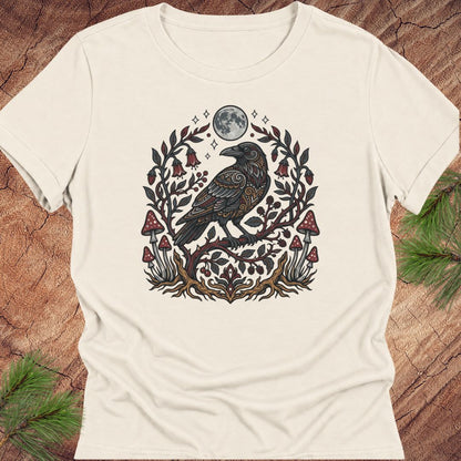 An off-white short-sleeve t-shirt with a detailed graphic of a black raven surrounded by a floral wreath, red mushrooms, and a full moon, displayed flat on a rustic wood background with pine needles.