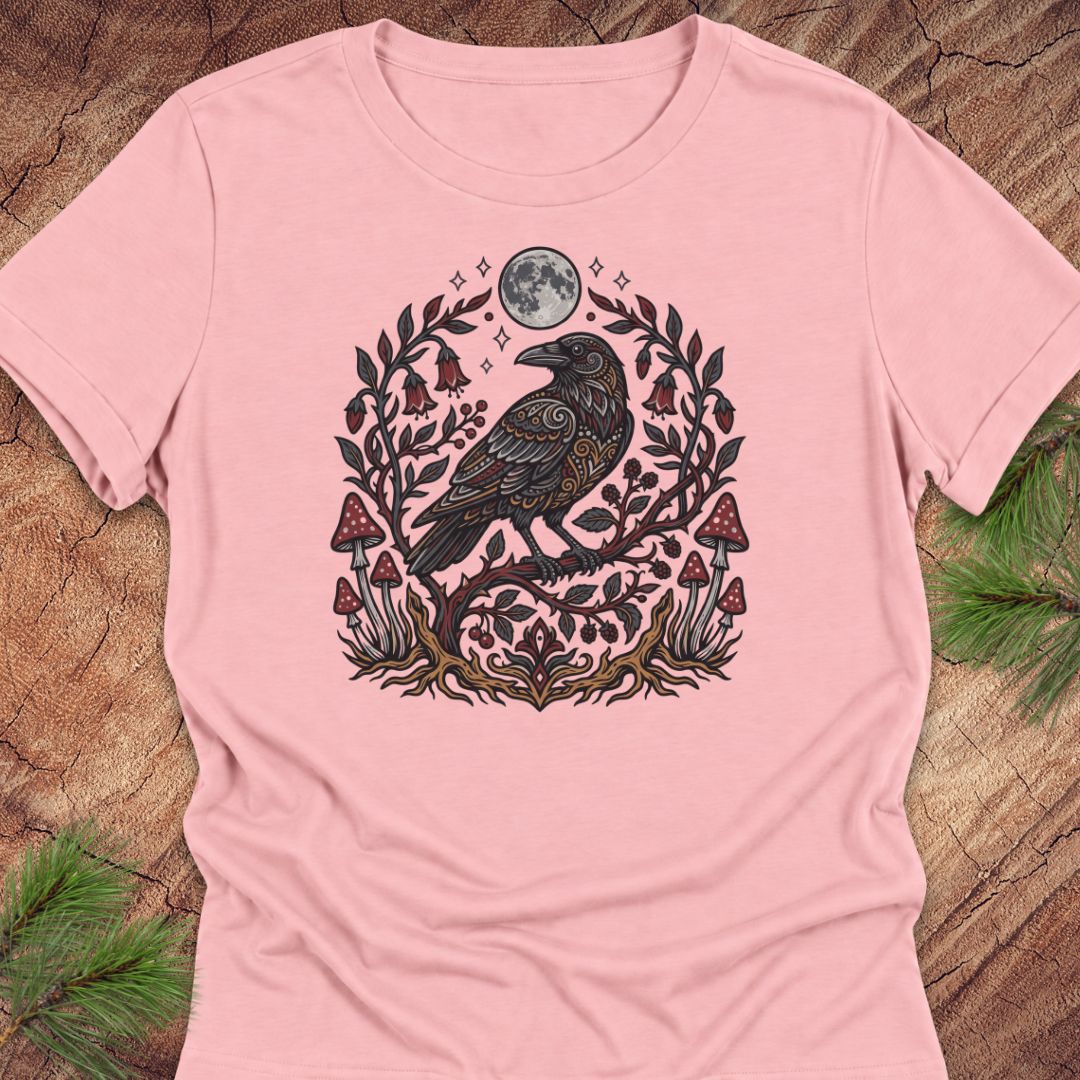 A light pink short-sleeve t-shirt with a detailed graphic of a black raven surrounded by a floral wreath, mushrooms, and a full moon, laid flat on a rustic wood background with pine needles.
