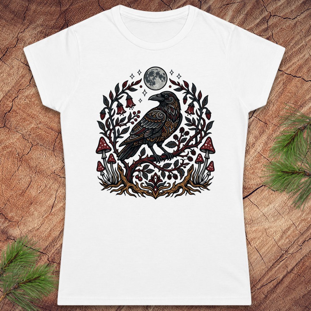 A white short-sleeve crewneck t-shirt featuring a detailed graphic of a black raven under a full moon, surrounded by a decorative wreath of leaves and red mushrooms, laid flat on a rustic wood background with pine branches.