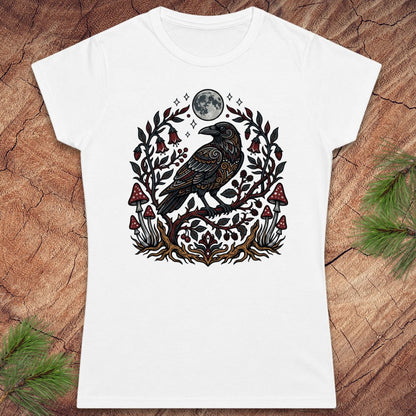A white short-sleeve crewneck t-shirt featuring a detailed graphic of a black raven under a full moon, surrounded by a decorative wreath of leaves and red mushrooms, laid flat on a rustic wood background with pine branches.