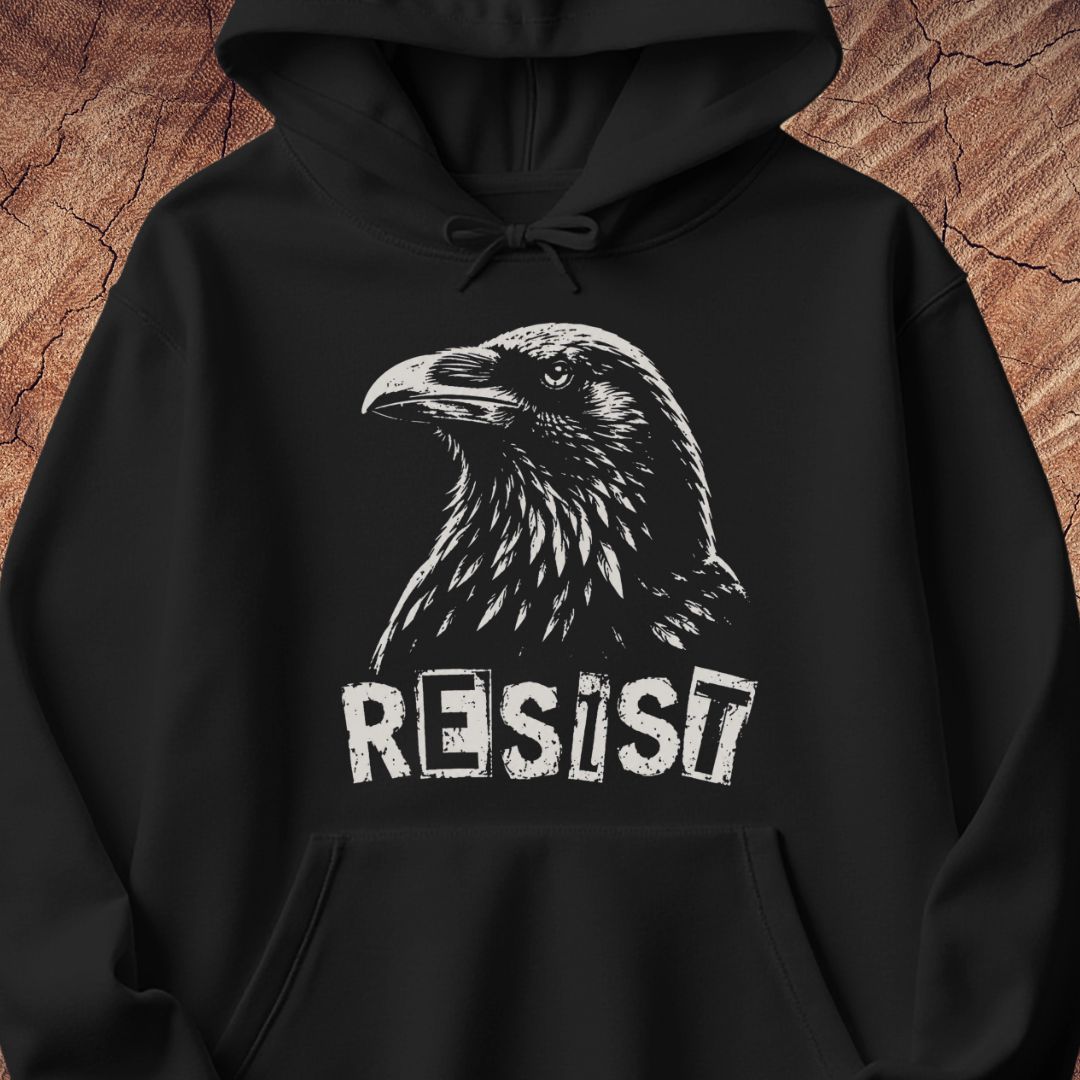 A black pullover hoodie with a white graphic print of a raven's head and the word 'RESIST' underneath, displayed against a textured brown wood background.