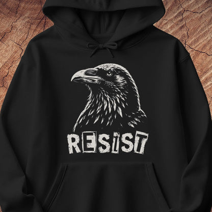 A black pullover hoodie with a white graphic print of a raven's head and the word 'RESIST' underneath, displayed against a textured brown wood background.