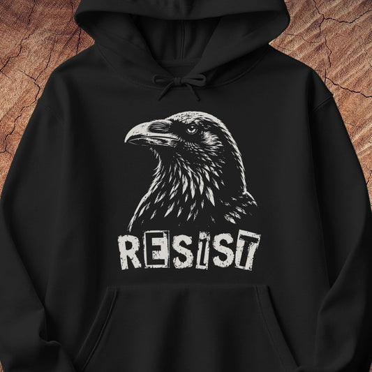 A black pullover hoodie with a white graphic print of a raven's head and the word 'RESIST' underneath, displayed against a textured brown wood background.