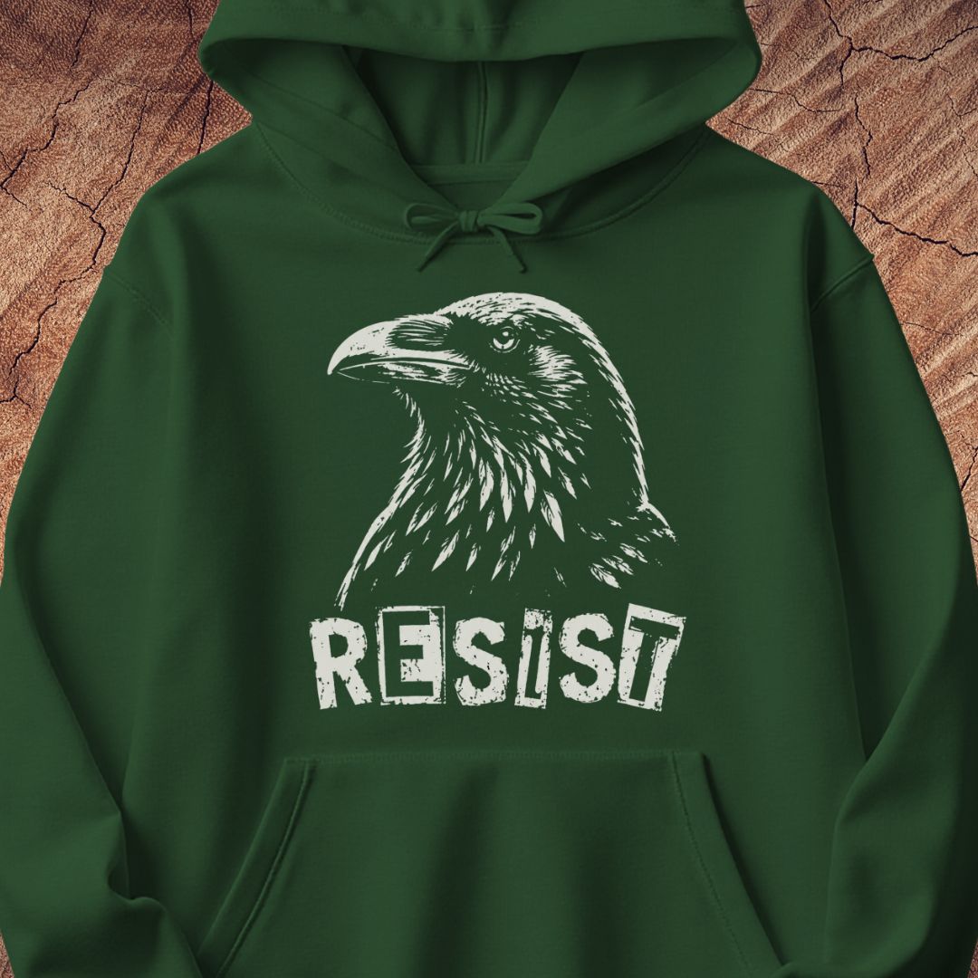 A dark green pullover hoodie featuring a white graphic print of a raven's head and the word 'RESIST' in a distressed stencil font, displayed against a textured brown wood background.