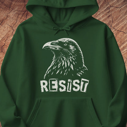 A dark green pullover hoodie featuring a white graphic print of a raven's head and the word 'RESIST' in a distressed stencil font, displayed against a textured brown wood background.