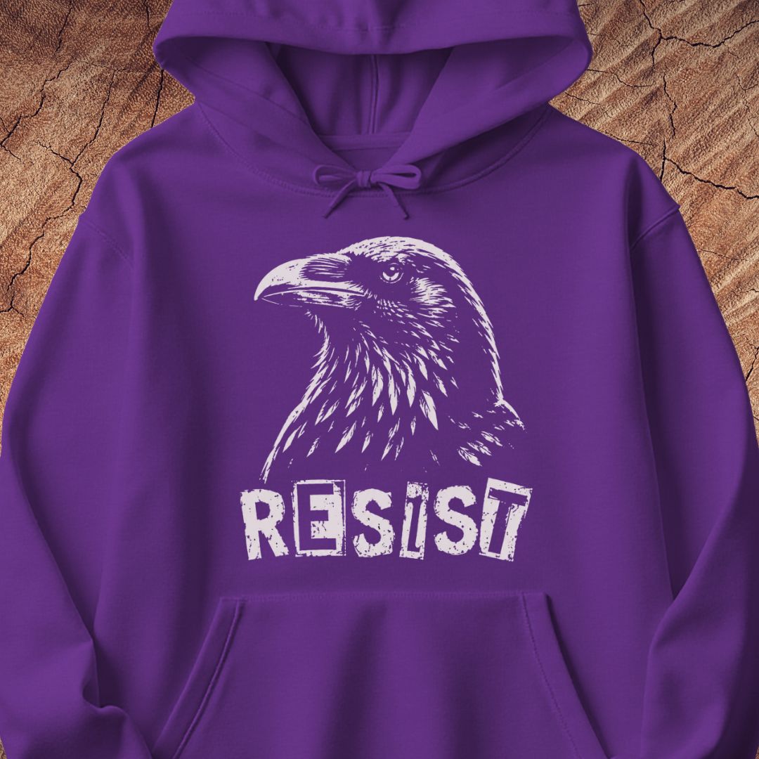 A purple pullover hoodie with a white graphic of a raven and the word 'RESIST' printed on the front, displayed against a rustic wood background.