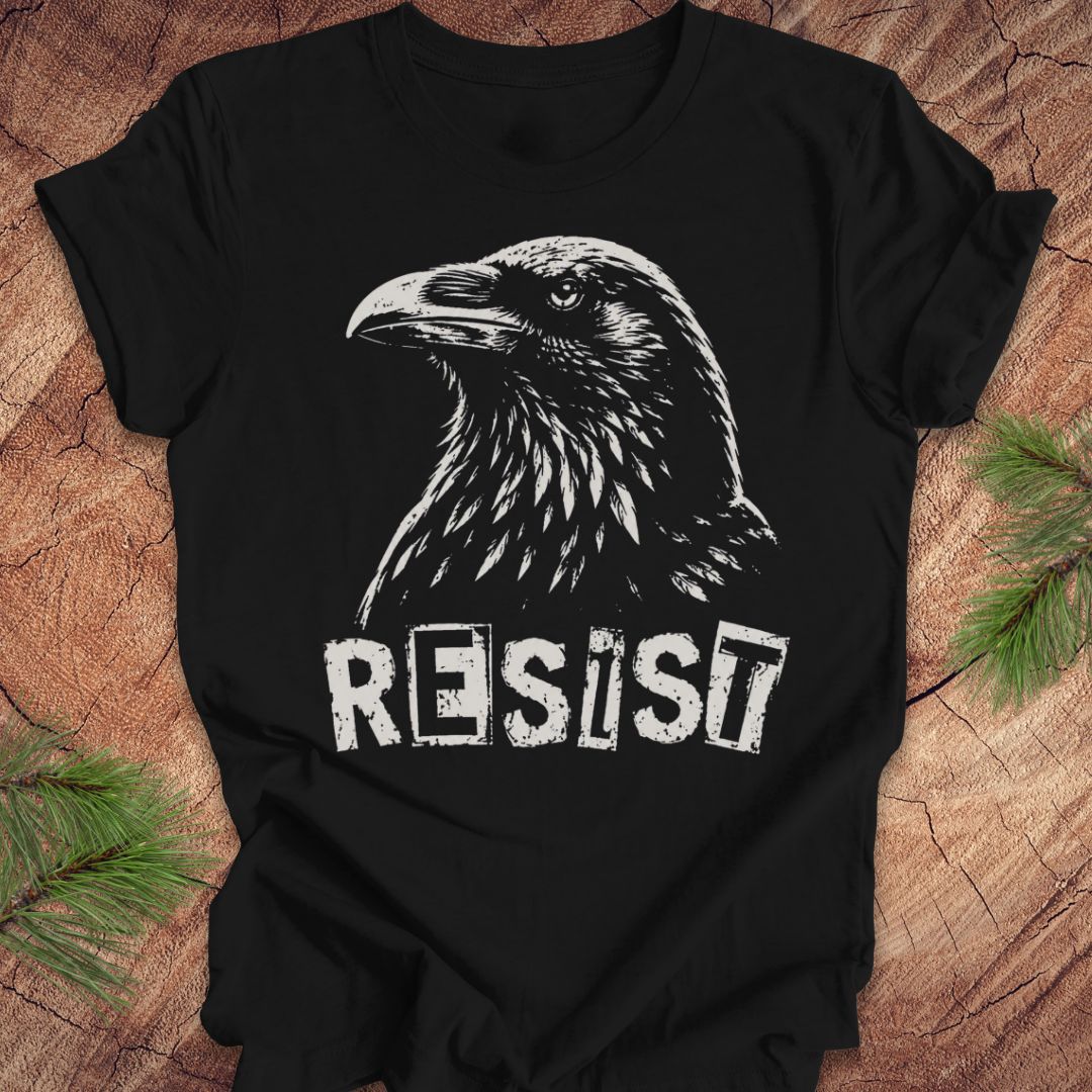 A black short-sleeve t-shirt with a white graphic of a raven and the word 'RESIST' printed on the front, laid flat on a rustic wood background with pine branches in the corners.