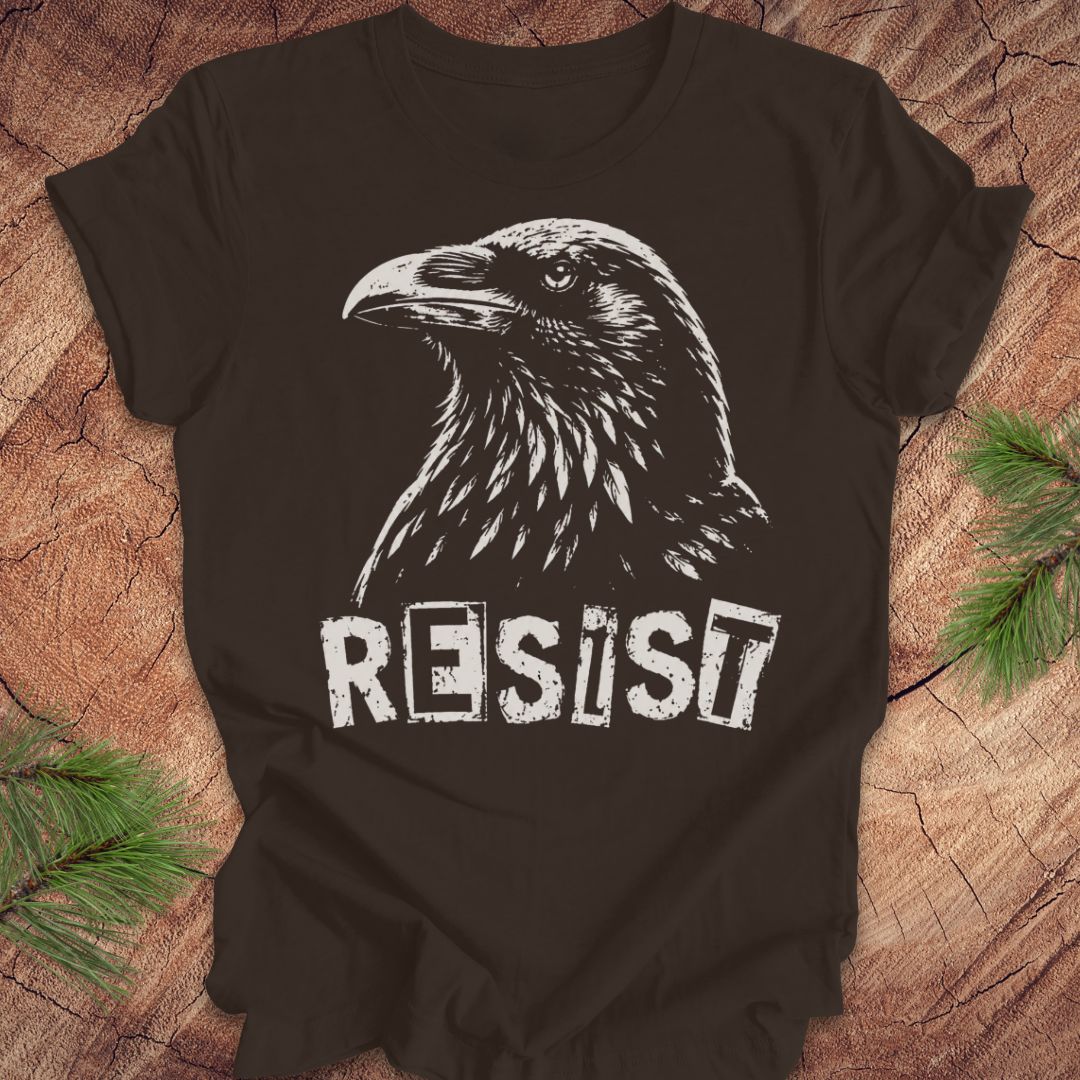 A brown short-sleeve t-shirt with a white graphic of a raven and the word 'RESIST' printed on the front, laid flat on a rustic wood background with pine branches in the corners.