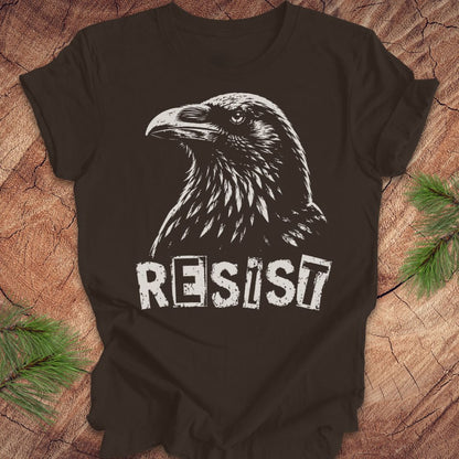 A brown short-sleeve t-shirt with a white graphic of a raven and the word 'RESIST' printed on the front, laid flat on a rustic wood background with pine branches in the corners.