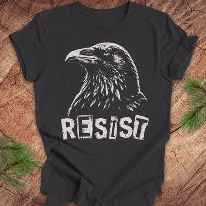 A dark gray short-sleeve t-shirt with a white graphic of a raven and the word 'RESIST' printed on the front, laid flat on a rustic wood background with pine branches in the corners.