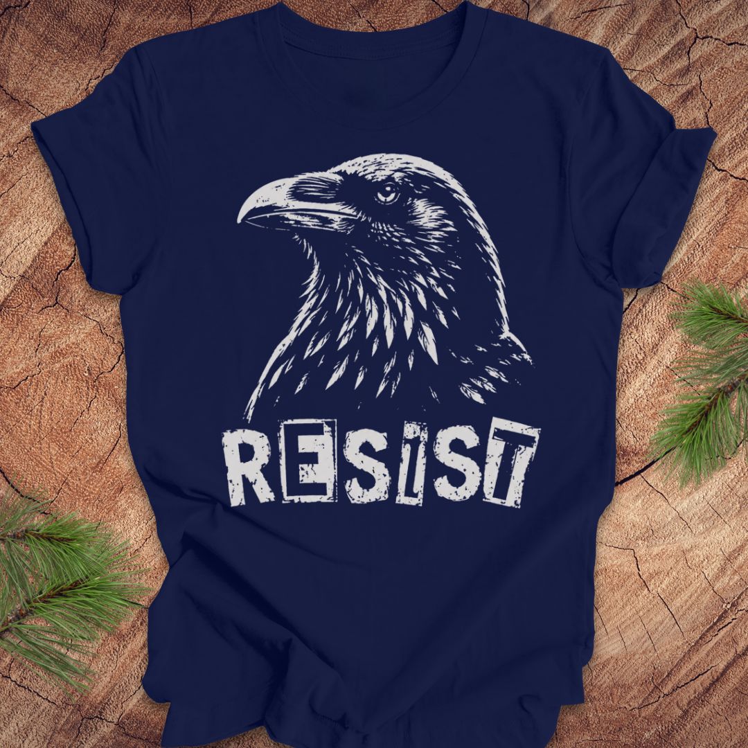 A navy blue short-sleeve t-shirt with a white graphic of a raven and the word 'RESIST' in a distressed stencil font, laid flat on a rustic wood background with pine branches in the corners.