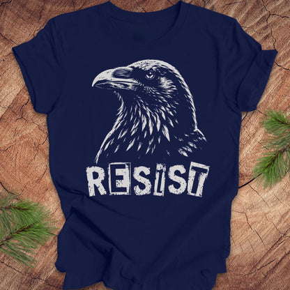A navy blue short-sleeve t-shirt with a white graphic of a raven and the word 'RESIST' in a distressed stencil font, laid flat on a rustic wood background with pine branches in the corners.