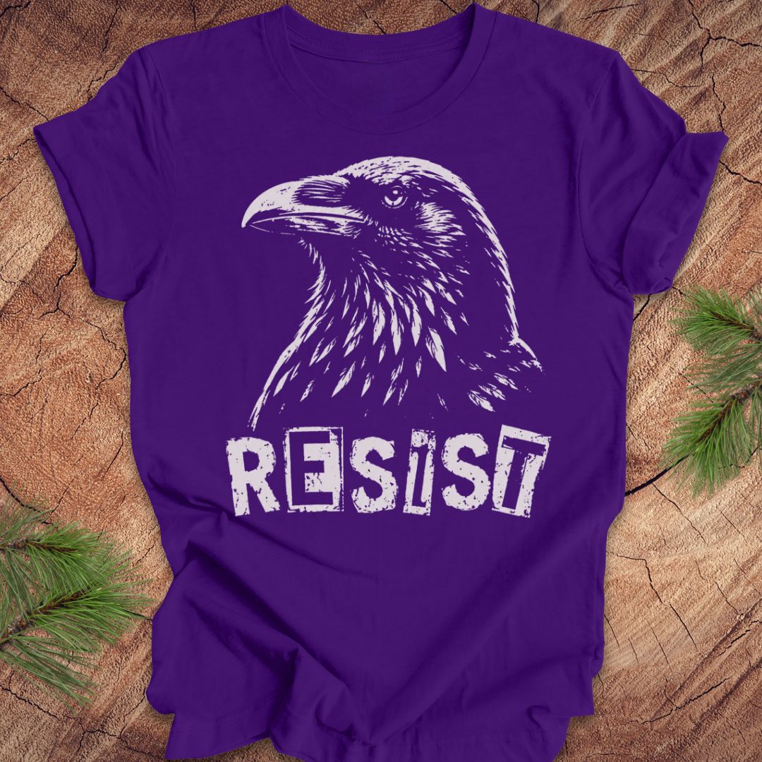 A purple short-sleeve t-shirt with a white graphic of a raven and the word 'RESIST' printed on the front, laid flat on a rustic wood background with pine branches in the corners.