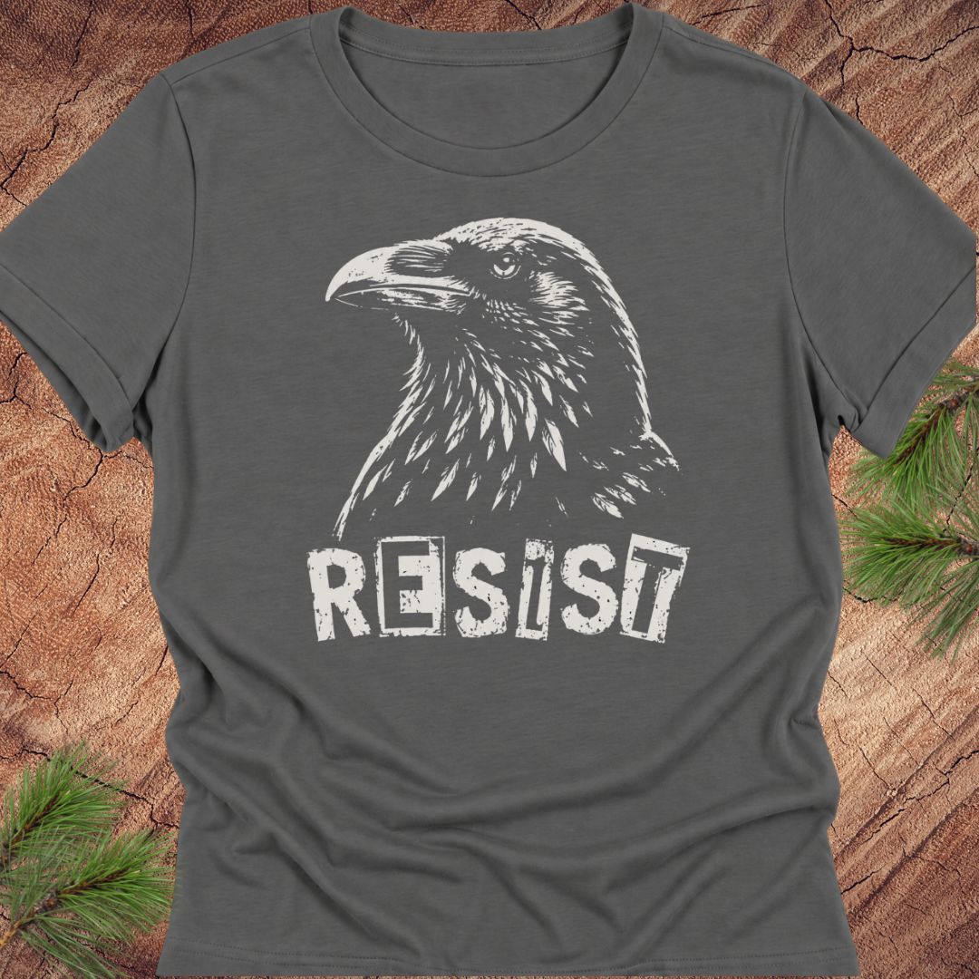 A dark gray crewneck t-shirt featuring a white graphic of a raven and the word 'RESIST' in a distressed stencil font, laid flat on a rustic wood background with pine needles in the corners.