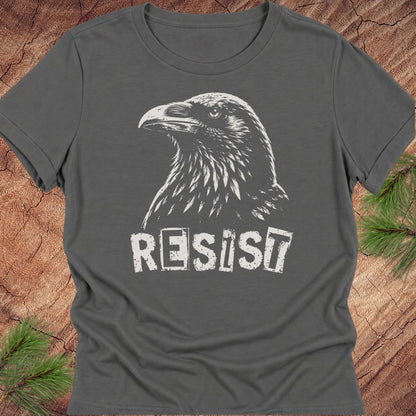 A dark gray crewneck t-shirt featuring a white graphic of a raven and the word 'RESIST' in a distressed stencil font, laid flat on a rustic wood background with pine needles in the corners.