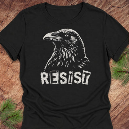 A black short-sleeve t-shirt with a white graphic of a raven and the word 'RESIST' printed on the front, laid flat on a rustic wood background with pine branches in the corners.