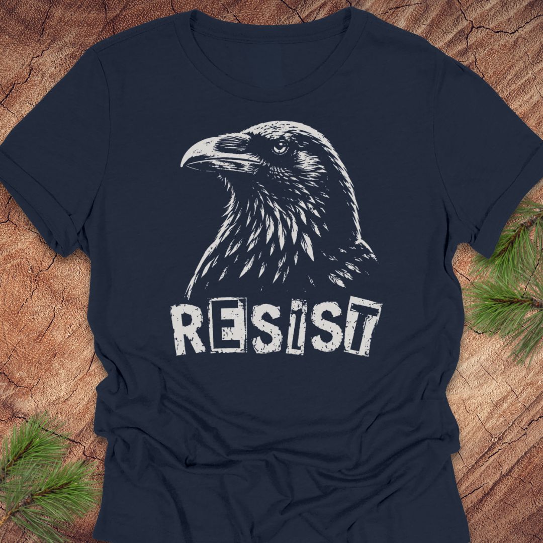 A navy blue short-sleeve t-shirt with a distressed white graphic of a raven and the word 'RESIST' printed on the front, laid flat on a rustic wood background with pine branches in the corners.