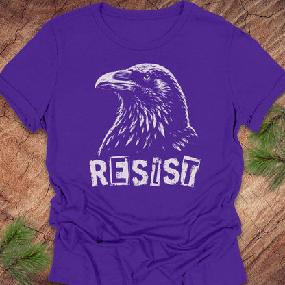 A purple short-sleeve t-shirt with a white graphic of a raven and the word 'RESIST' printed on the front, laid flat on a rustic wood background with pine branches in the corners.