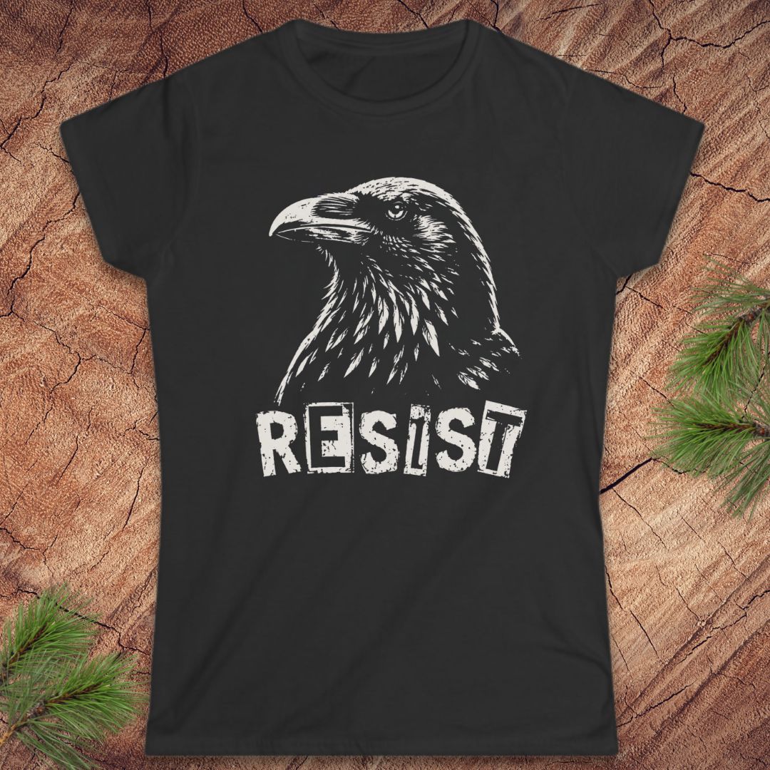 A black short-sleeve t-shirt with a white graphic of a raven and the word 'RESIST' printed on the front, displayed flat on a rustic wood background with pine branches in the corners.