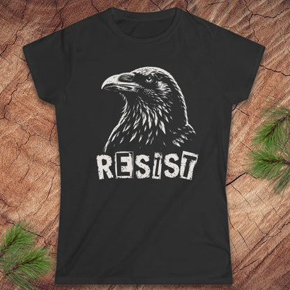 A black short-sleeve t-shirt with a white graphic of a raven and the word 'RESIST' printed on the front, displayed flat on a rustic wood background with pine branches in the corners.