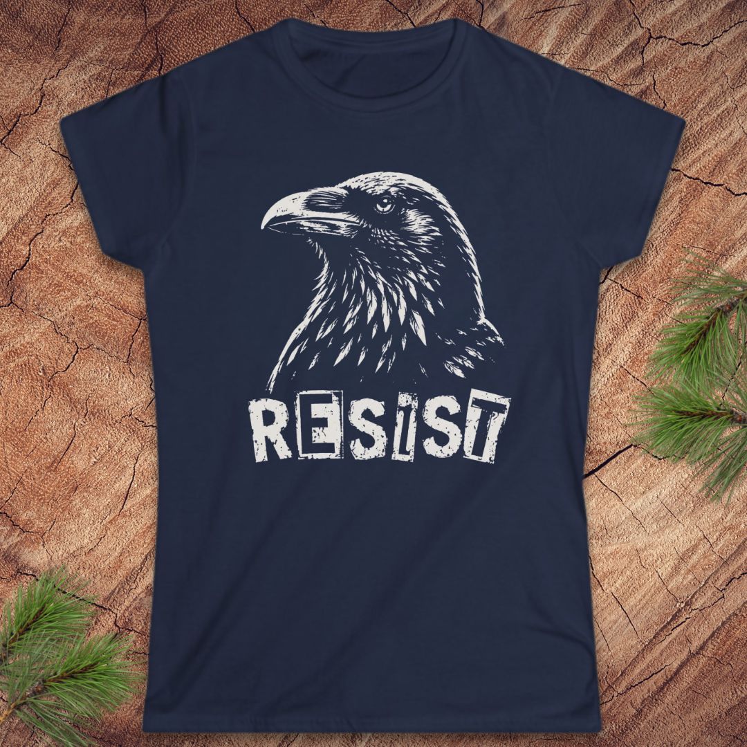 A navy blue short-sleeve t-shirt with a white graphic of a raven and the word 'RESIST' printed on the front, displayed flat on a rustic wood background with pine branches in the corners.