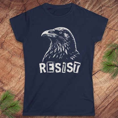 A navy blue short-sleeve t-shirt with a white graphic of a raven and the word 'RESIST' printed on the front, displayed flat on a rustic wood background with pine branches in the corners.