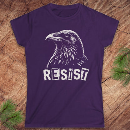 A purple short-sleeve t-shirt with a white graphic of a raven and the word 'RESIST' printed on the front, displayed flat on a wood background with pine branches in the corners.