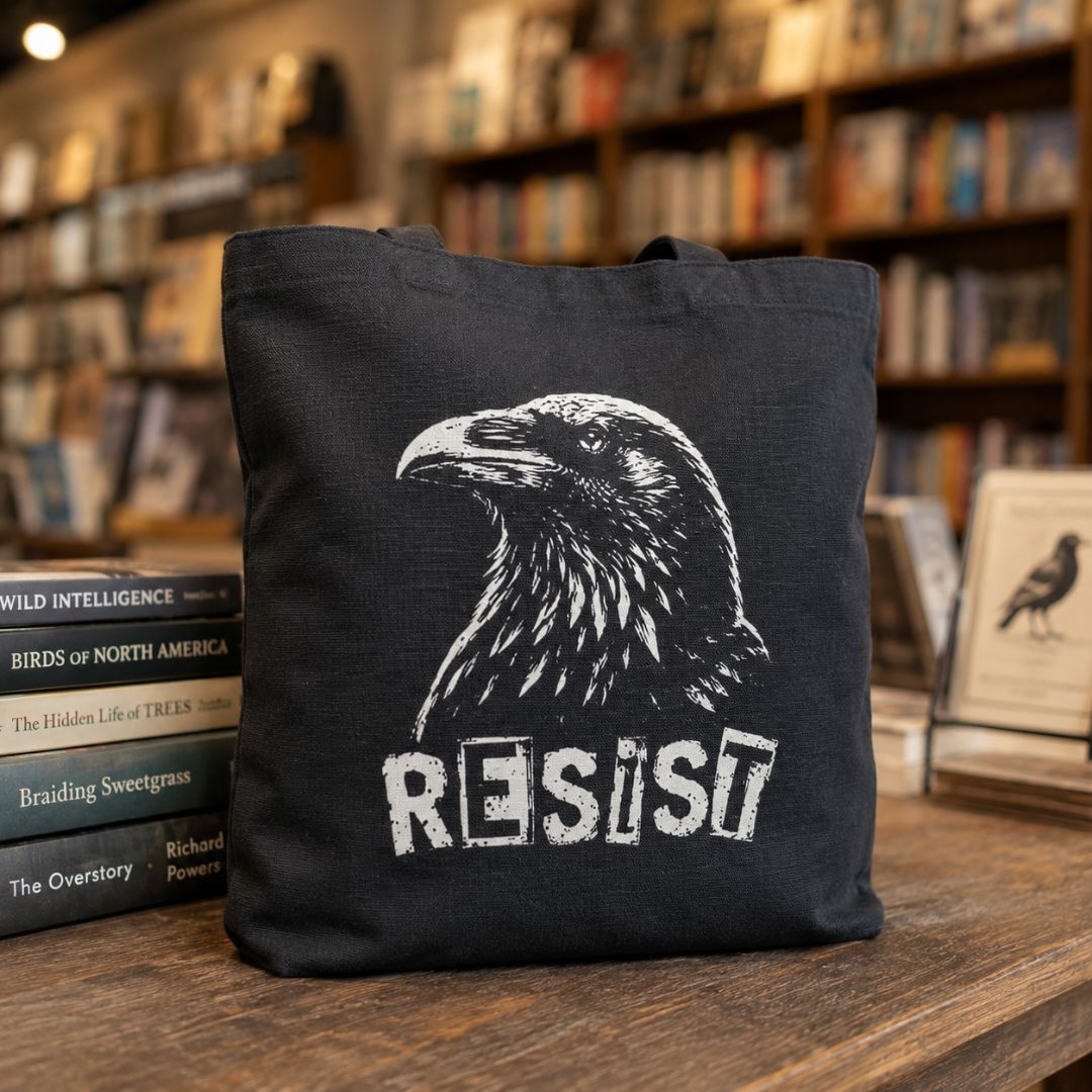 A black canvas tote bag with a white graphic of a raven and the word 'RESIST' printed on the front, displayed on a wooden table in a bookstore setting.