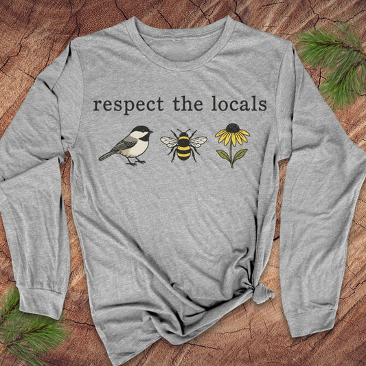 A heather gray long-sleeve t-shirt with a graphic print reading 'respect the locals' above a chickadee, a bee, and a yellow flower. The shirt is styled flat on a rustic wood background with pine needles.
