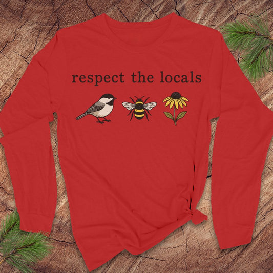 A red long-sleeve t-shirt with a graphic print reading 'respect the locals' above a chickadee, a bee, and a yellow flower. The shirt is displayed flat on a rustic wood background with pine branches in the corners.