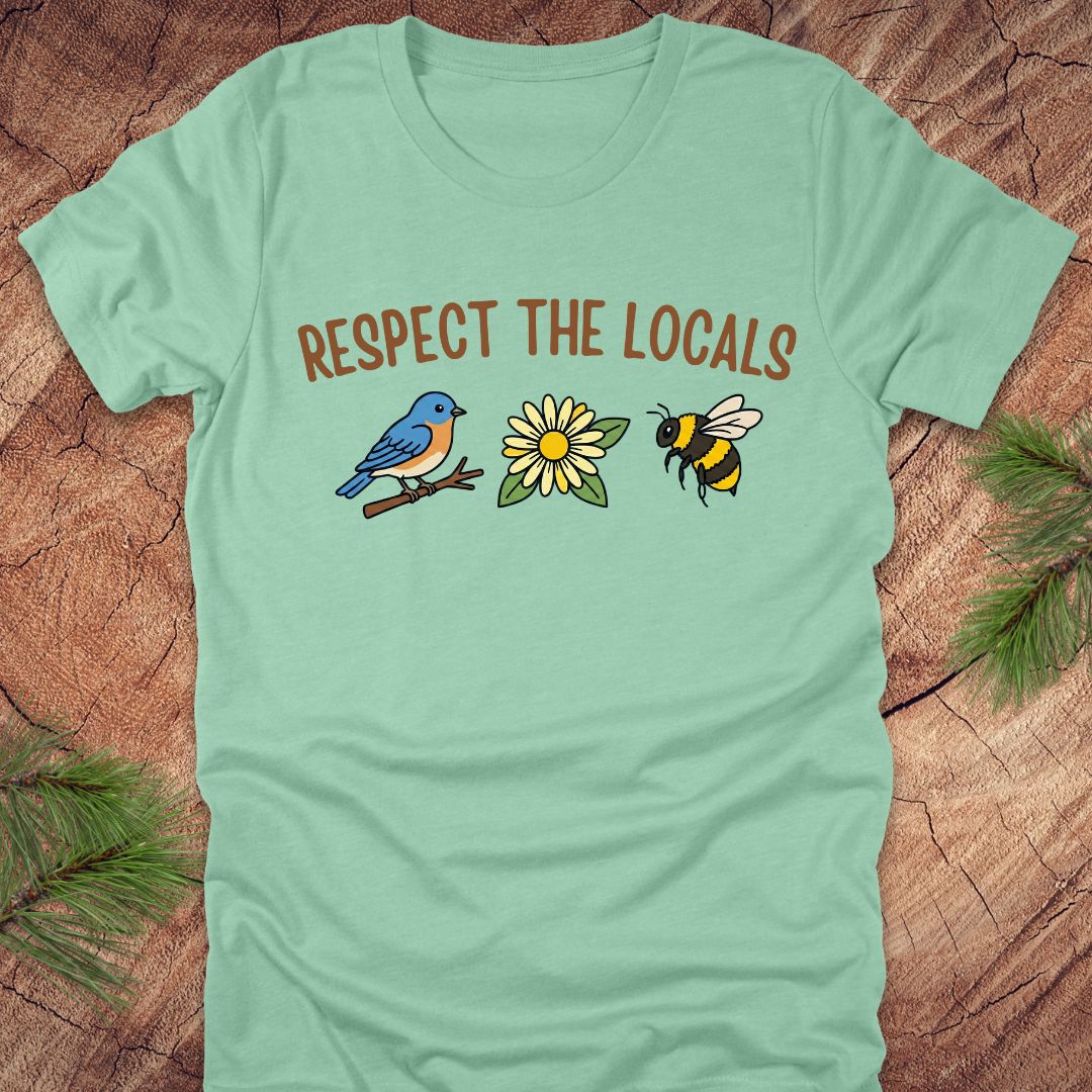 Mint green t-shirt with 'Respect the Locals' text and bird, flower and bee graphics on a wooden surface