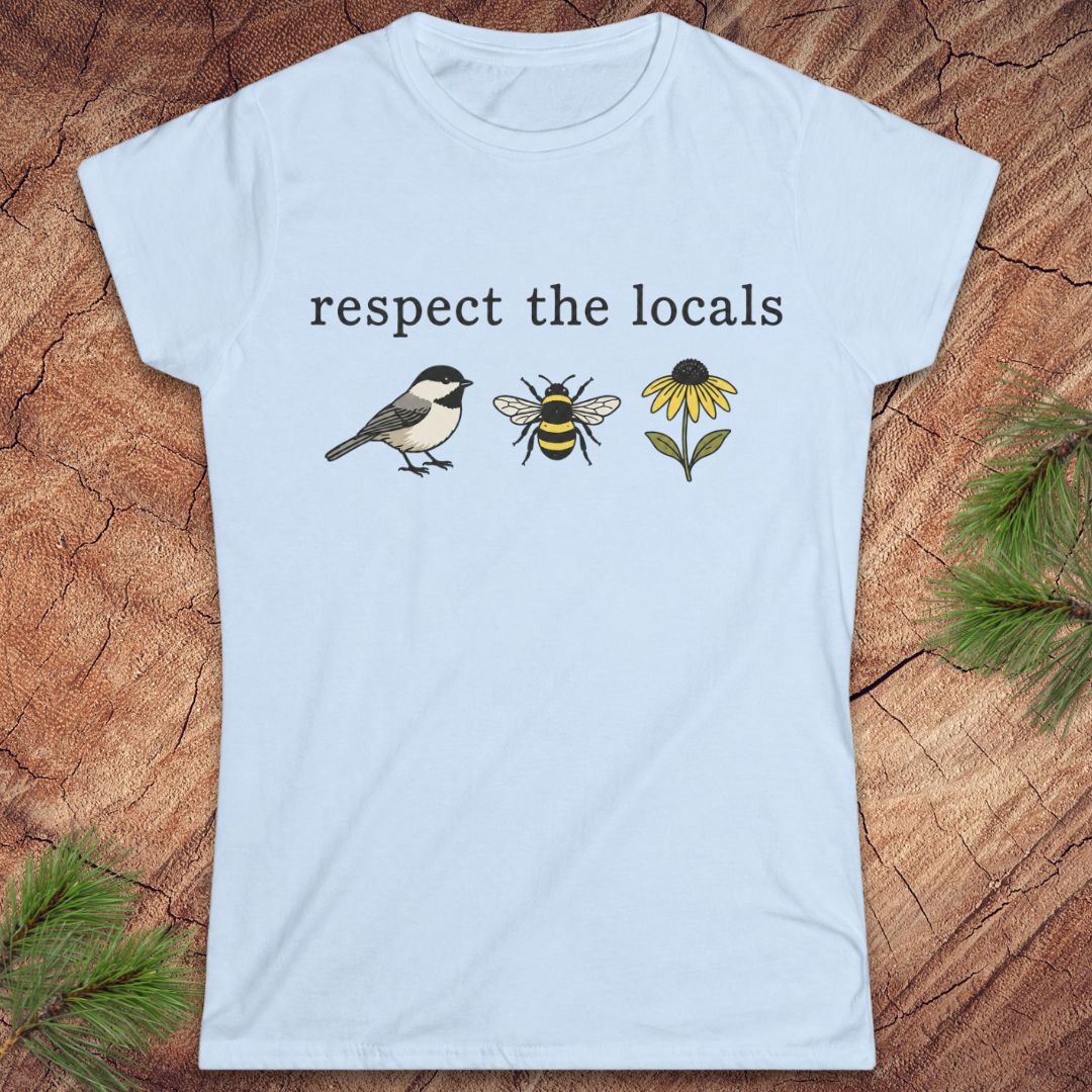 A light blue short-sleeve t-shirt with a graphic print on the front reading 'respect the locals' above a bird, a bee, and a yellow flower. The shirt is displayed flat on a rustic wood background with pine branches in the corners.