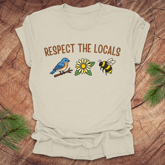 Beige t-shirt with 'Respect the Locals' text and bird, flower and bee graphics on a wooden surface