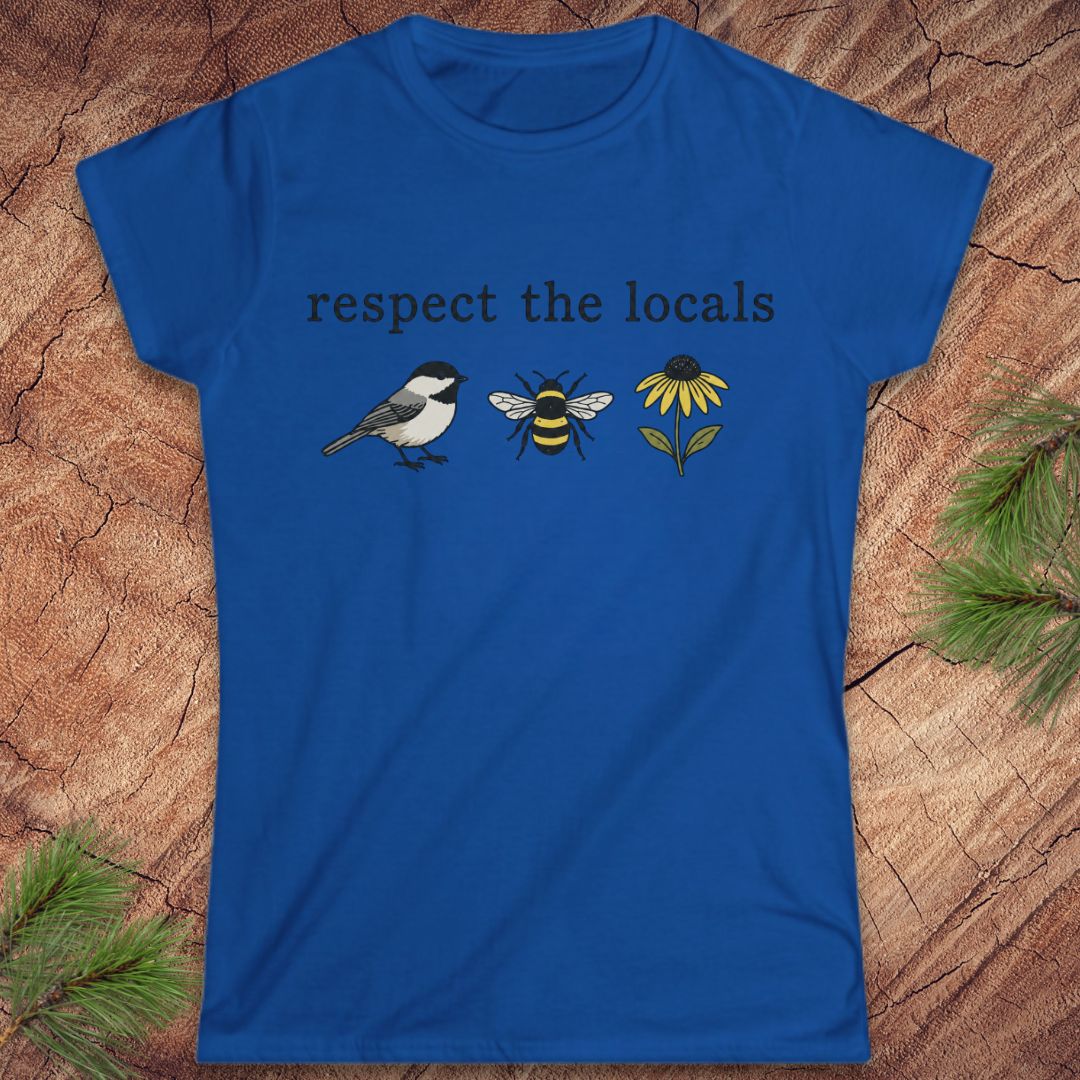 A royal blue short-sleeve t-shirt with a graphic print reading 'respect the locals' above a bird, a bee, and a yellow flower. The shirt is laid flat on a textured wood background with pine branches in the corners.