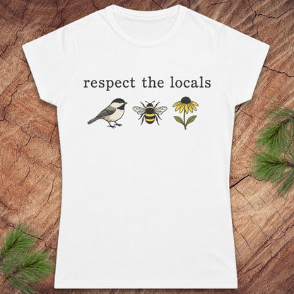 A white short-sleeve crewneck t-shirt with a graphic print on the front reading 'respect the locals' above a chickadee, a bee, and a yellow flower. The shirt is displayed flat on a textured brown wood background with pine needles in the corners.