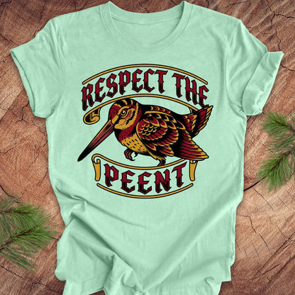 Light green t-shirt with a colorful graphic of a bird and text 'Respect the Peent' on a wooden surface.