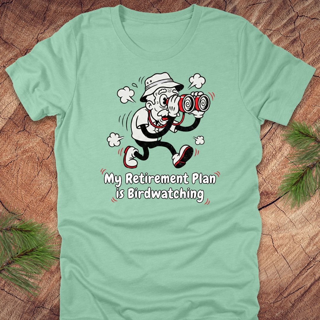 Light green t-shirt with a cartoon character and text 'My Retirement Plan is Birdwatching' on a wooden surface.