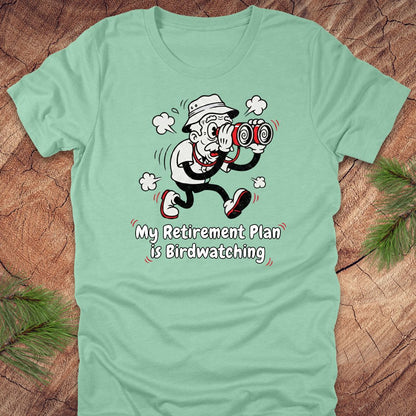 Light green t-shirt with a cartoon character and text 'My Retirement Plan is Birdwatching' on a wooden surface.