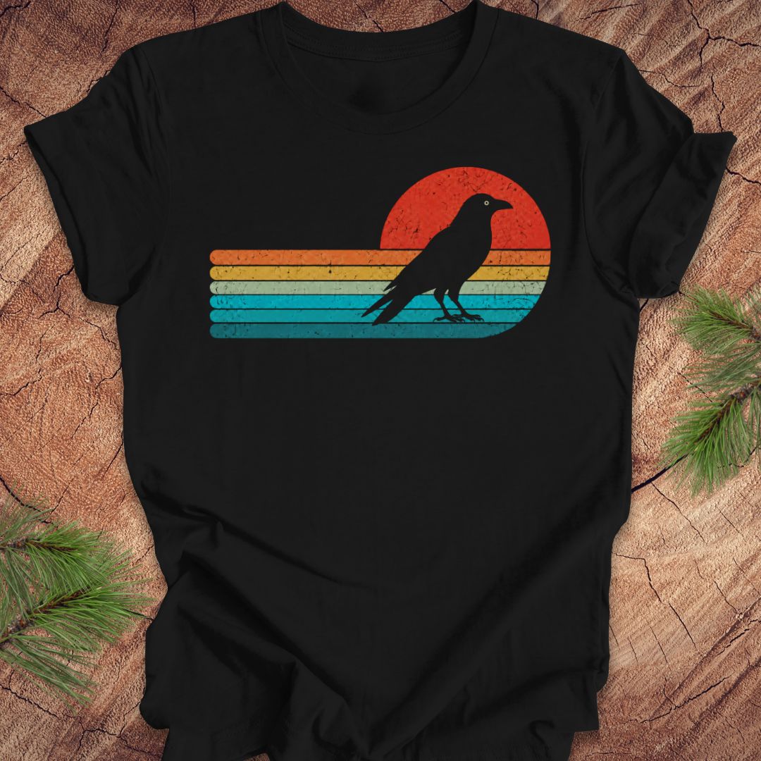 A black short-sleeve t-shirt laid flat on a rustic wood surface, featuring a distressed retro sunset graphic with horizontal stripes in orange, yellow, and blue, and a black raven silhouette standing in front of the sun.